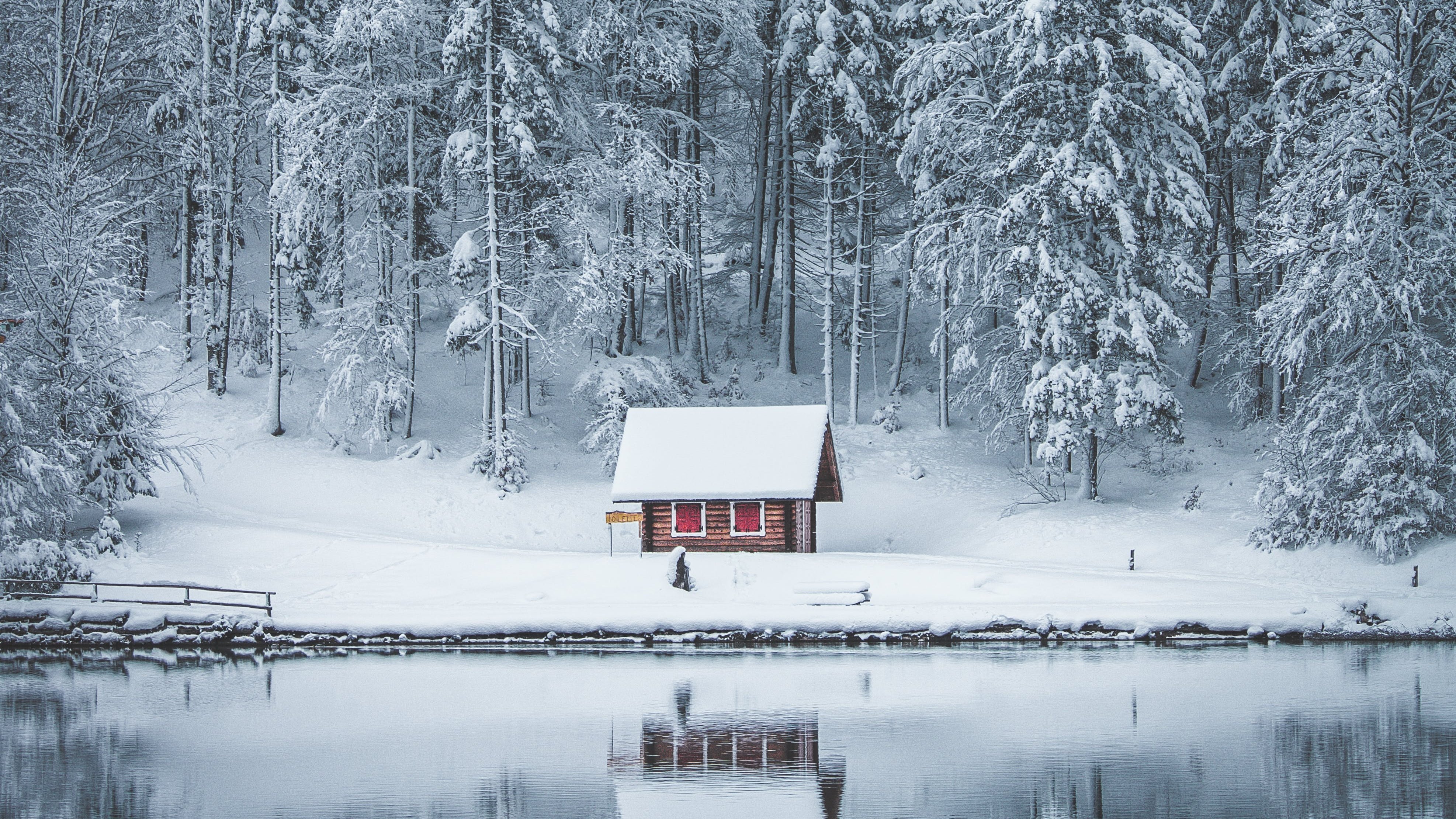 Kleine Hütte mit roten Fensterläden an einem See, im Hintergrund verschneiter Nadelwald